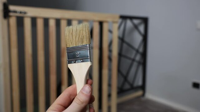 A hand holds a 2 inch natural bristle paintbrush. A wooden gate with vertical slats and a black geometric metal frame is visible in the indoor setting.