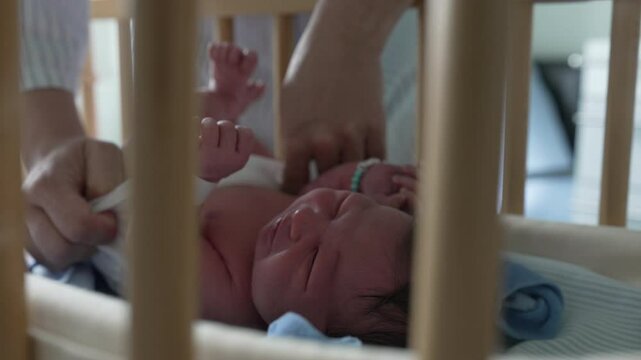 Mother&rsquo;s hands carefully adjusting newborn baby&rsquo;s clothing in a bassinet, viewed through crib bars, highlighting the tender and loving care provided in early postpartum