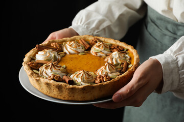 Woman holding homemade pumpkin pie with whipped cream, seeds and cinnamon on black background, closeup