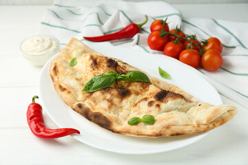 Tasty calzone, basil, chili pepper, sauce and tomatoes on white wooden table, closeup