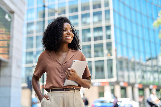 Happy confident young African American professional woman, student, female entrepreneur, businesswoman looking away holding tab standing on business city street dreaming of future career. Copy space.