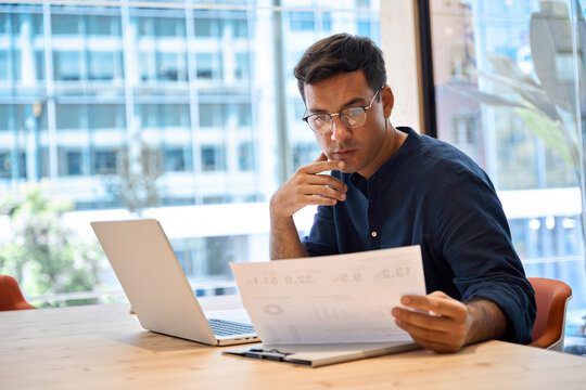Busy young business man employee using laptop computer technology checking financial document working in office. Professional businessman executive, company worker or manager looking at paper at work.