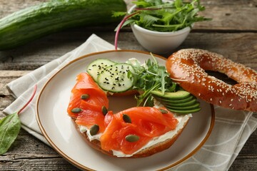 Delicious bagel with salmon, cream cheese, cucumber, avocado and pumpkin seeds on wooden table, closeup