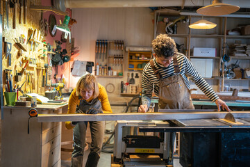 Two partners joiners operate woodworking machine, cutting lumber for production and measuring timbers. Carpenters learning to cut with precision, skilled mentoring and commitment to family business