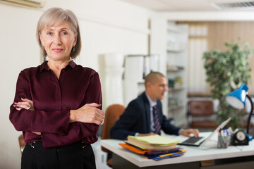 Senior woman office worker standing and looking in camera. Man in suit sitting in background.