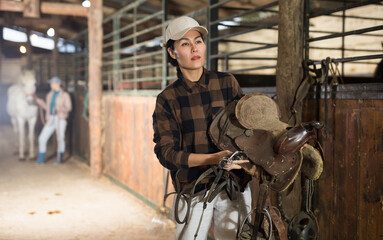 Confident smiling Asian female stable keeper carrying horse saddle and harness for horseback ride