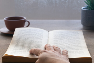 Hand turning the page of an open book on a wooden table, in the background a window with unfocused...