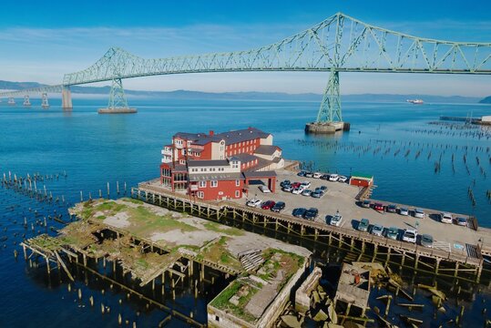 Aerial view of the Cannery Pier Hotel & Spa, a red building on a pier, with cars parked below. A large green bridge spans the water. Uniontown, Astoria, Oregon, USA