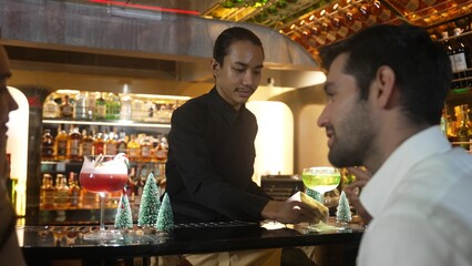 Special winter Midori Sour cocktail with caramelized citrus slice served by bartender serving smart guest talking couple with punch cocktail with candy cane christmas theme at festive bar. Vinosity.
