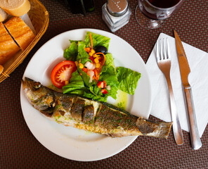 There is plate with fried whole carcass zander fish and mix vegetable salad on table. Grated carrot, corn, china cabbage, olive, corn and tomato complement fried fish