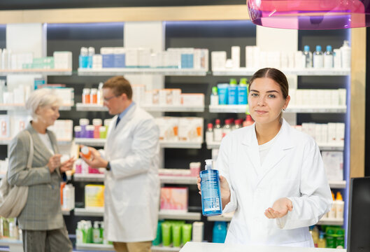 Polite young female pharmacist demonstrating lubricant in chemist's shop with large assortment of products