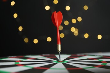 Red arrow hitting target on dart board against black background with blurred lights, closeup