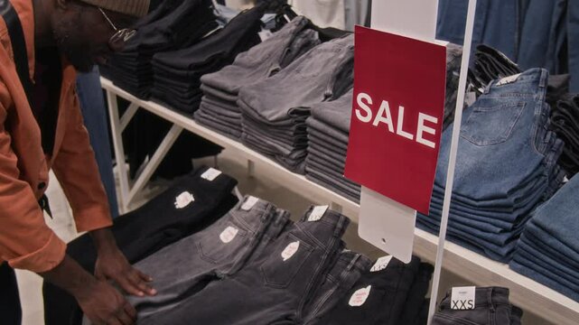 Medium shot of African American man in casual clothes choosing jeans in sales section of clothing store