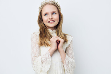 Young girl with a delicate crown, smiling in elegant white dress against a light pastel background...