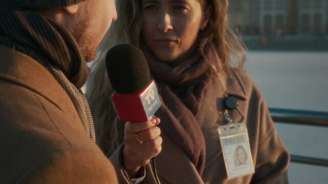 Medium closeup of unrecognizable young man in smart coat speaking to female TV reporter in city street, who is listening and holding news channel microphone, while conducting public opinion poll