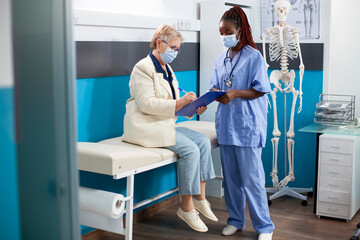 Obraz premium In medical office, retired white woman fills out forms with guidance from female healthcare worker. African american nurse patiently waits as elderly patient writes medical information on clipboard.