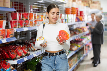 Young woman in supermarket buys instant fast food in bowl. Girl buyer reads information on packaging of semi-finished product. Concept of affordable box-container feeding