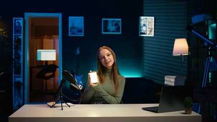 Cheerful gen Z girl in home studio reviewing mini RGB lights controller, using microphone to...