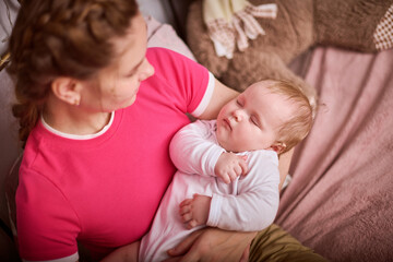 Young woman with braided hair gently cradles sleeping baby girl in pink and beige room. Warm lighting and soft textures create calm atmosphere