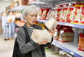 In imported goods store, senior woman buyer examines and view product, Chinese noodles. Mature female customer gets acquainted with unconventional and unfamiliar funchuosa product in store.