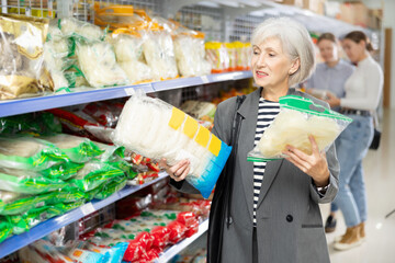 Mature female in shop carefully examines information about product on label packaging. Elderly woman customer chooses and buys pack of Chinese noodles at hypermarket