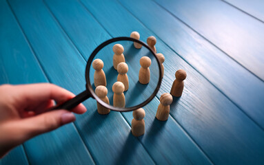 A hand holds a magnifying glass over a group of wooden figures, focusing on a few in the center, while others remain blurry in the background.