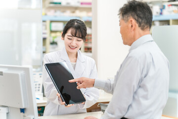 Obraz premium 薬局でタブレットPCを使いながら説明する薬剤師・調剤師（レセコン・レセプトコンピュータ） 