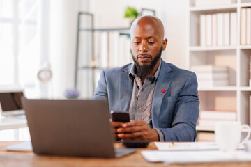 Obraz premium African American Businessman using mobile phone while working with laptop at workplace office. browsing site on cellphone.