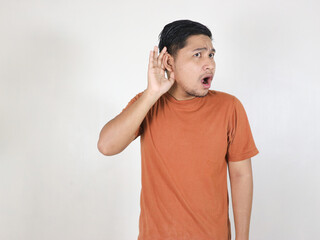 asian man wearing brown t-shirt standing on white background is eavesdropping and showing shocked...