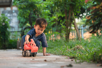 Thai child in denim outfit playing outdoors with a red toy truck, engaged in imaginative play on a paved garden path.