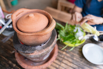 Clay pot on a traditional charcoal stove, used for Thai hotpot cooking with fresh vegetables nearby.