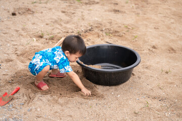 Young Thai child in a floral outfit playing with sand near a plastic tub, engaging in outdoor exploration and creativity.