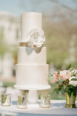 Elegant white wedding cake adorned with a decorative flower and surrounded by floral arrangements on a sunny day