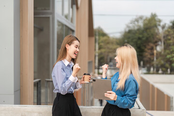 Diverse businesswomen celebrating success while discussing ideas outdoors with a tablet and coffee.