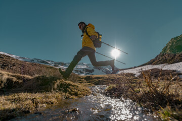 A man in bright clothes with a backpack and trekking poles jumps over a stream against a backdrop of snowy mountains and the sun.