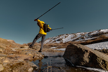 A tourist in bright clothes and carrying a backpack, holding trekking poles, jumps from a rock to the ground. There are snowy mountains and clear sky on the background.