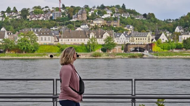 Following along next to a woman walking next to the Rhine River with an old small town on a hill in the distance, Koblenz, Rhine Valley, Germany