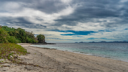 A secluded sandy beach on a tropical island. A calm turquoise ocean. Green vegetation on the shore. A picturesque cliff in the distance. Mountains on the horizon. Blue sky, clouds. Madagascar.