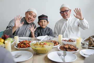 Portrait Of Multi Generation Muslim Family Greeting By Waving Hands At Dining Room Celebrating Eid Al Fitri 