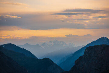 Deep gorge between layered silhouettes of sharp rocky ridge spurs under huge snowy mountain peak in golden cloudy sky. Sheer crags of mount valley under giant ice top under gold sunset color clouds.