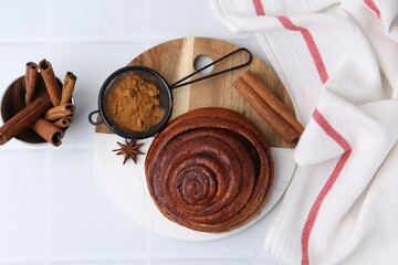 Delicious cinnamon roll bun and spices on white tiled table, flat lay