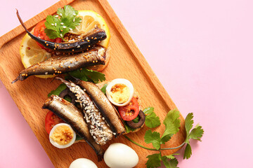 Wooden board of tasty sandwiches with canned smoked sprats on pink background