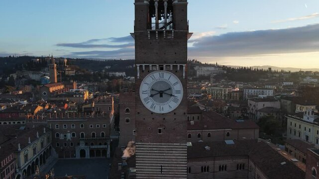 A breathtaking aerial backward shot starting from the clock of Torre dei Lamberti, Verona, at dawn, with the soft morning light gradually illuminating the tower and surrounding cityscape.