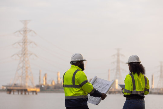Two engineers in safety gear review construction blueprints near a power plant and transmission towers. They appear to be conducting a site inspection along a river or waterfront facility.