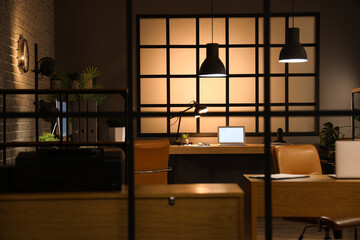 Interior of dark office with desks and glowing lamps at night, view through folding screen