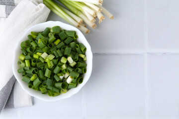 Chopped green onion in bowl and stems on white tiled table, top view. Space for text
