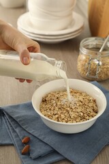 Woman pouring milk into bowl with oatmeal at wooden table, closeup