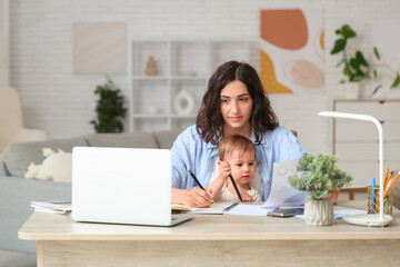 Cute little baby and young mother working with laptop while writing in notebook on maternity leave at home