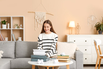 Happy female writer with typewriter, books and crumpled paper sitting on sofa at home