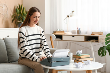 Beautiful happy female writer with typewriter, books and crumpled paper sitting on sofa at home